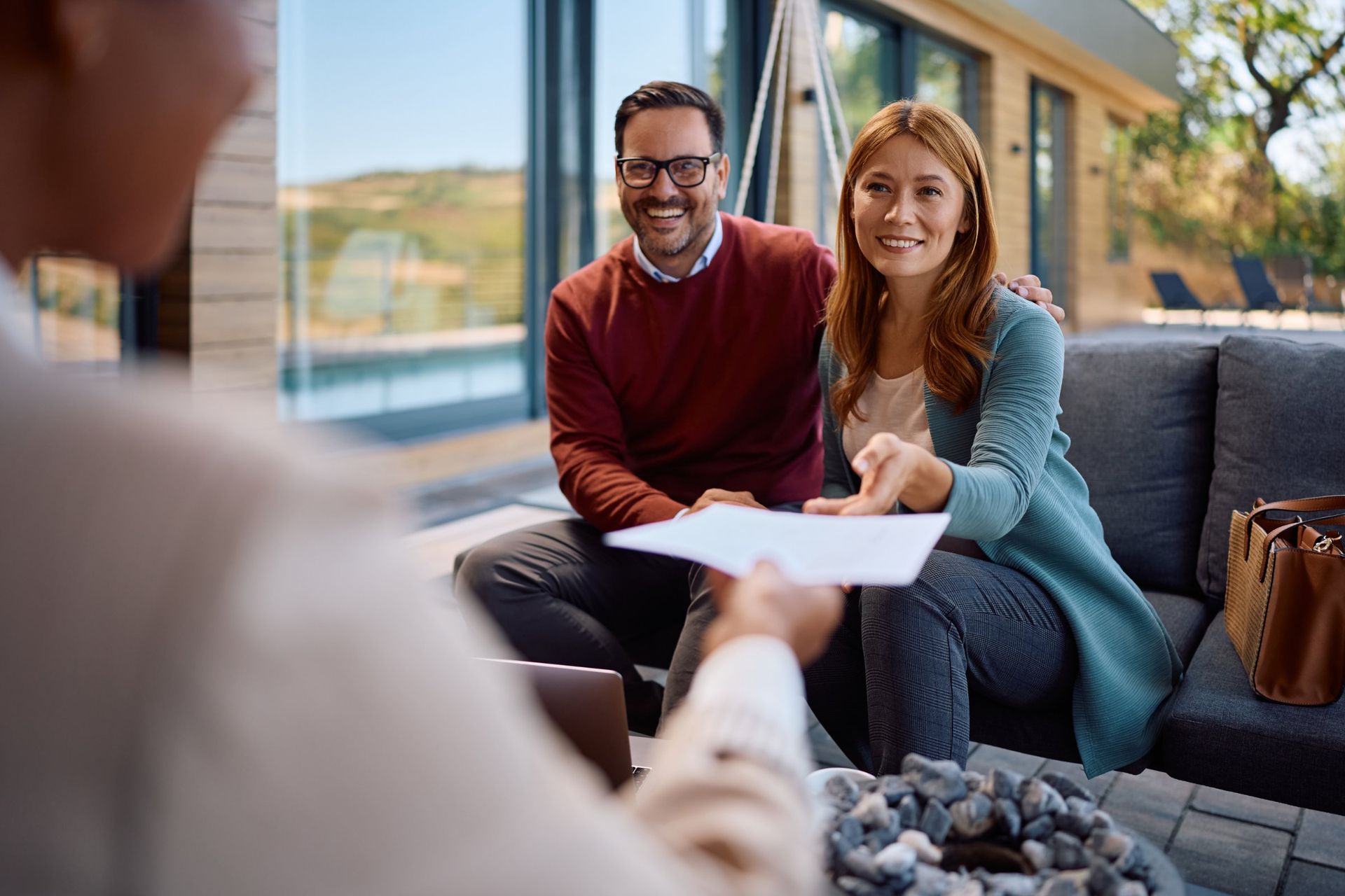 Couple smiles, accepting documents from a person.  They sit on a couch in a modern home, outdoors visible.