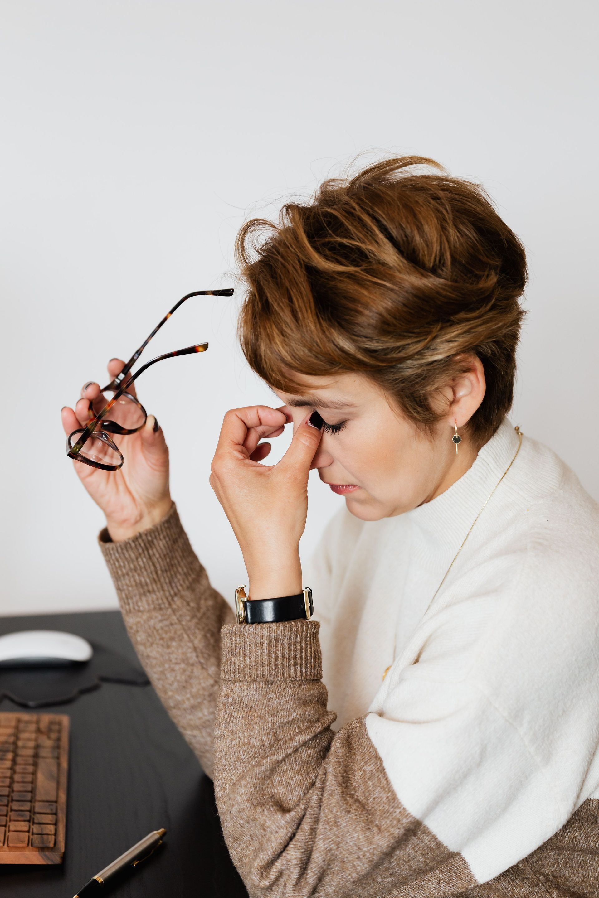 Erschöpfte Frau am Tisch, Brille in der Hand, Augen geschlossen.