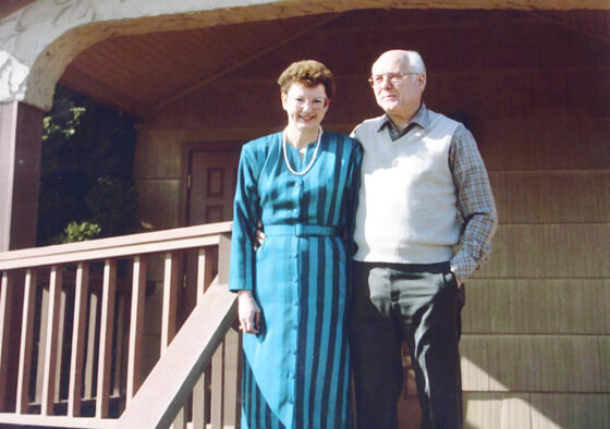 Couple standing on porch steps in front of a house. The woman wears a blue dress, the man a sweater vest.