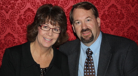 Woman with glasses and man in suit smiling in front of a red patterned backdrop.