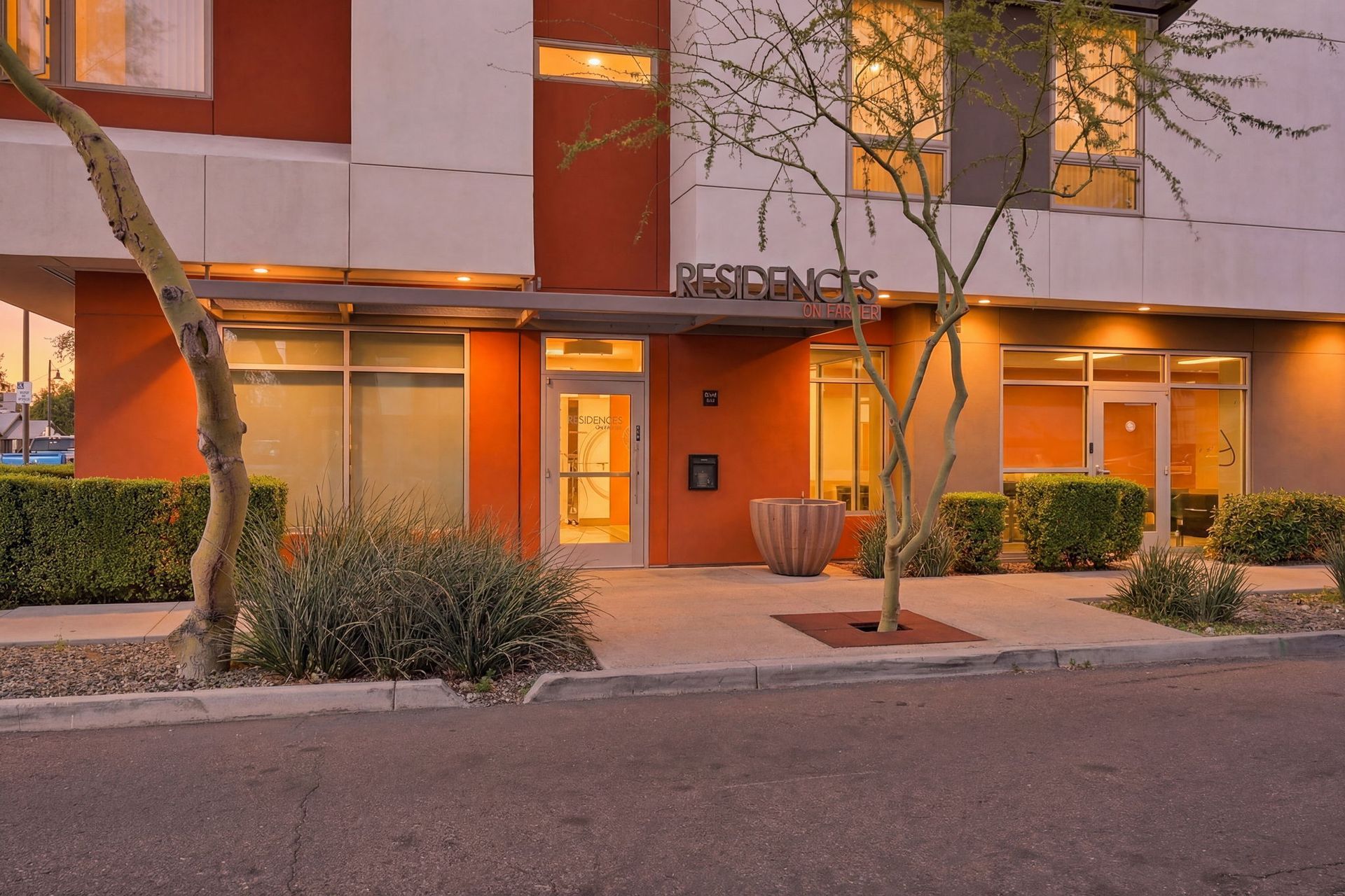 Orange apartment building entrance at dusk with lit doorways, trees, and shrubs along the sidewalk
