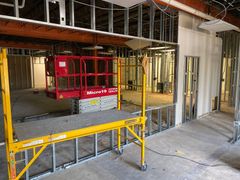 Indoor construction site with a red scissor lift behind yellow safety railings and exposed wall framing