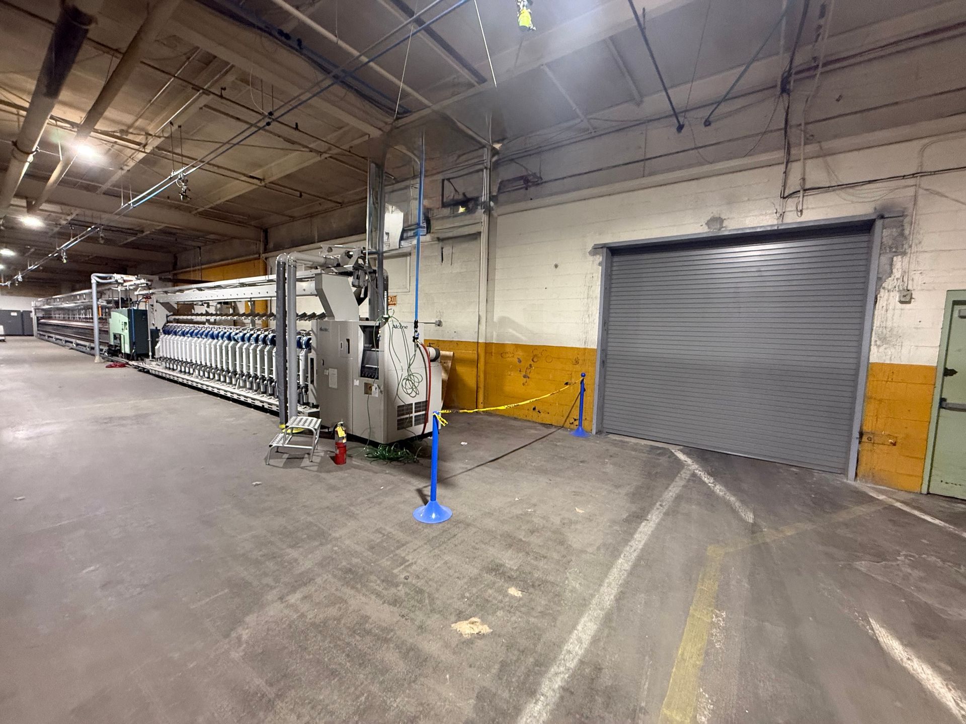 Industrial warehouse interior with a gray roll-up door, machinery, and stacked materials along the left wall.