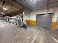 Industrial warehouse interior with a gray roll-up door, machinery, and stacked materials along the left wall.
