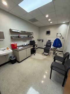 Medical exam room with exam chair, sink, shelves, cabinets, and waiting chairs under fluorescent lights