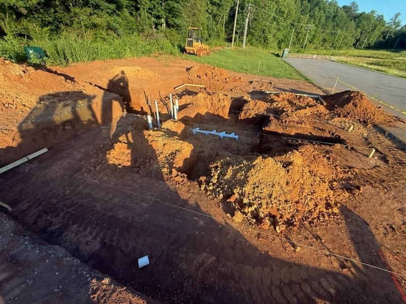 Large muddy excavation beside a road, with a person and a white dog near exposed pipes at sunset