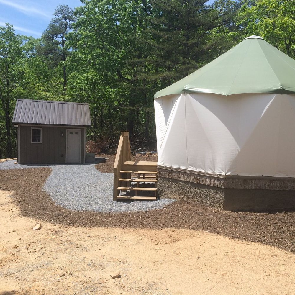 Round white yurt with green roof beside a small shed and gravel path in a wooded campground