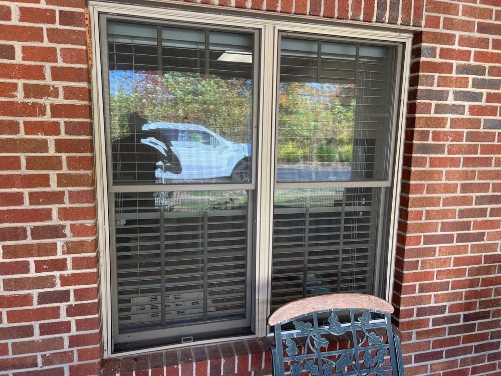 Brick exterior wall with a double window, white blinds, and a small metal chair in front