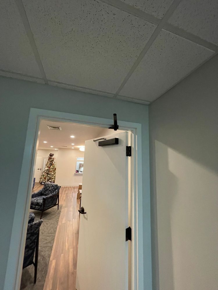 Open office doorway under a drop ceiling, showing a lit room with wood flooring and a decorated tree inside.