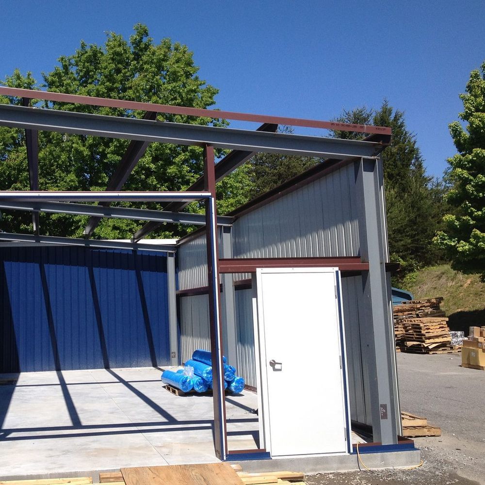Metal shed frame beside a white door and blue tarp in a gravel yard, with trees and pallets nearby