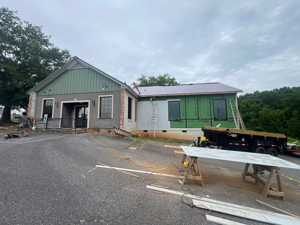 Small building with green trim beside a wet parking lot and construction materials, under overcast skies