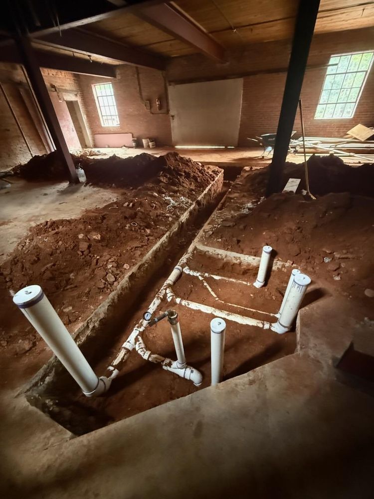Exposed plumbing pipes and dirt trenches inside a dim room under renovation