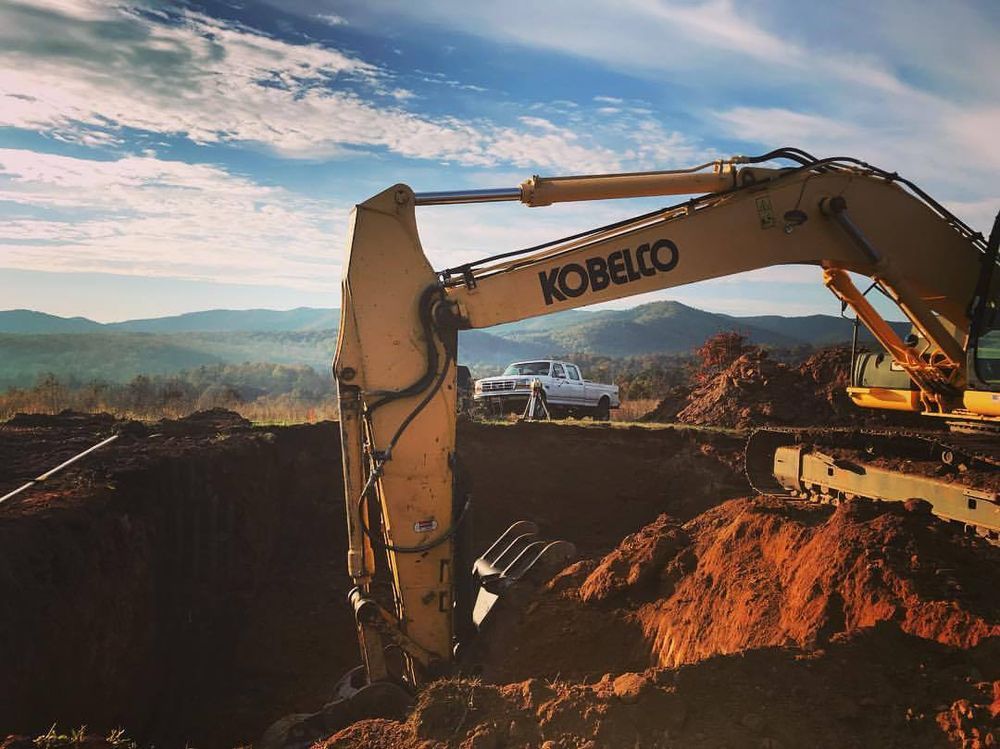 Excavator digging a large red dirt pit with mountains and blue sky in the background