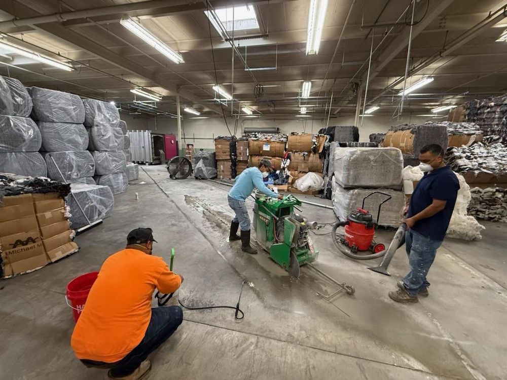 Workers operating machinery in a warehouse with stacked wrapped pallets and concrete floor
