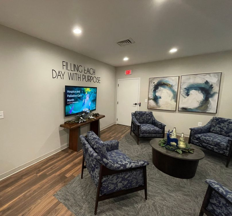 Small modern lounge with blue chairs, round table, TV, and wall art under “LIVING EACH DAY WITH PURPOSE” sign
