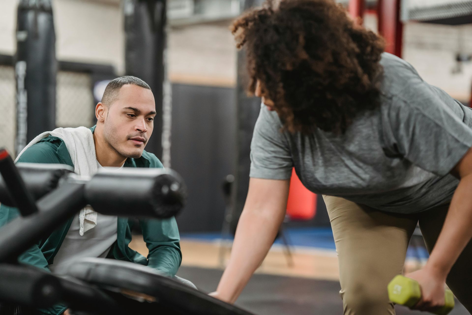 A person lifting a dumbbell while being supervised by another in a gym setting.