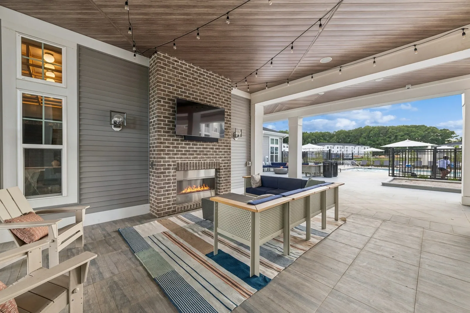 Covered outdoor lounge with brick fireplace, mounted TV, string lights, and pool beyond.