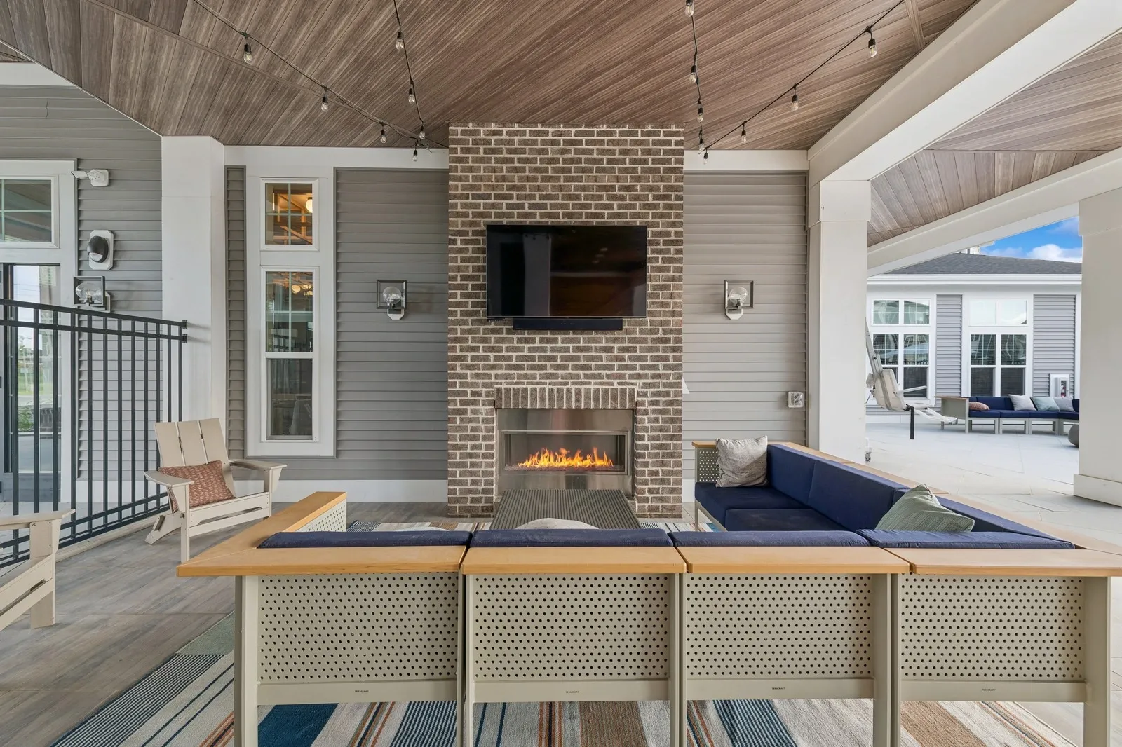 Outdoor covered lounge with brick fireplace, mounted TV, string lights, and blue sectional seating.