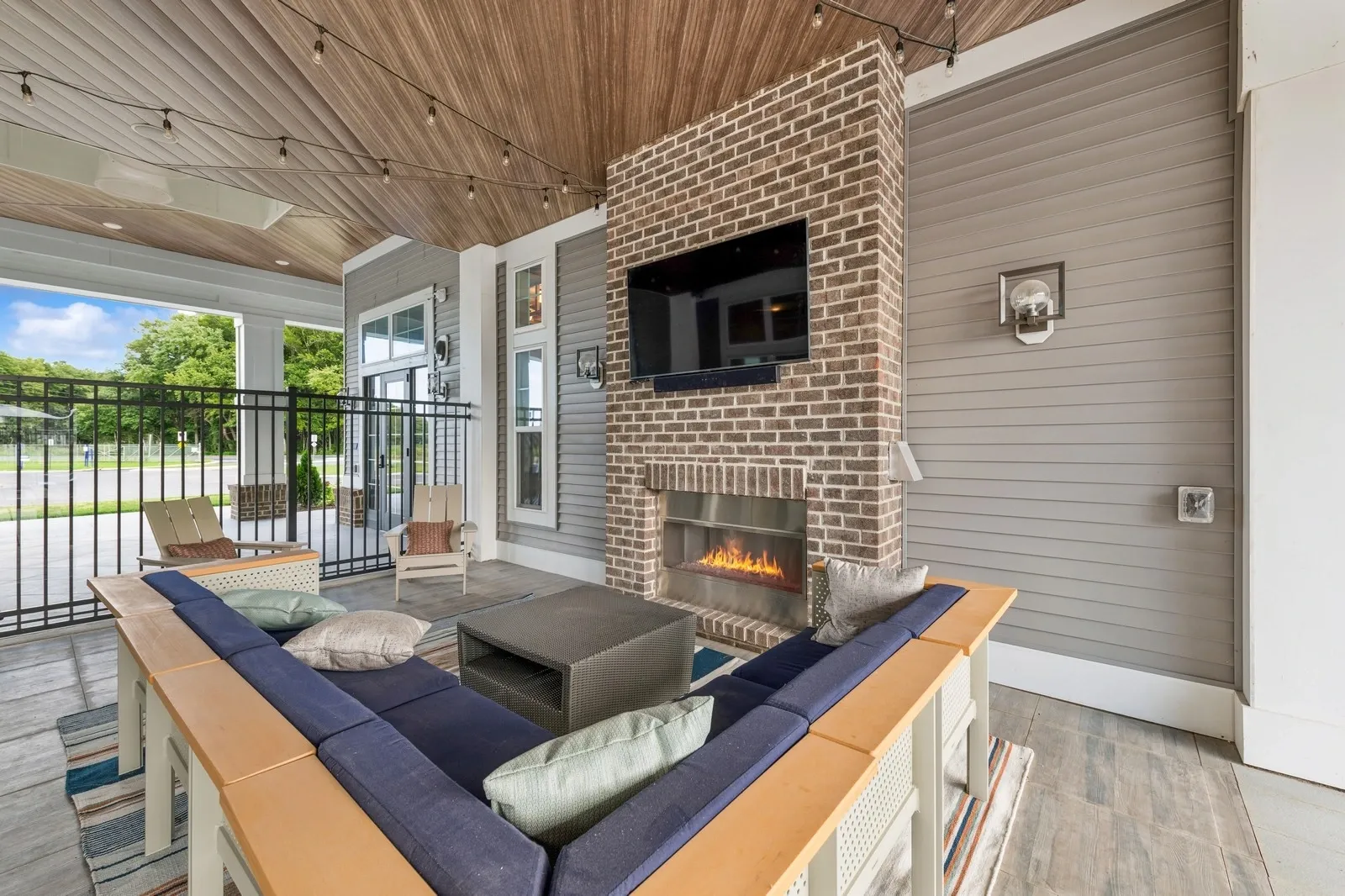 Outdoor covered lounge with brick fireplace, wall-mounted TV, string lights and blue-cushioned seati…