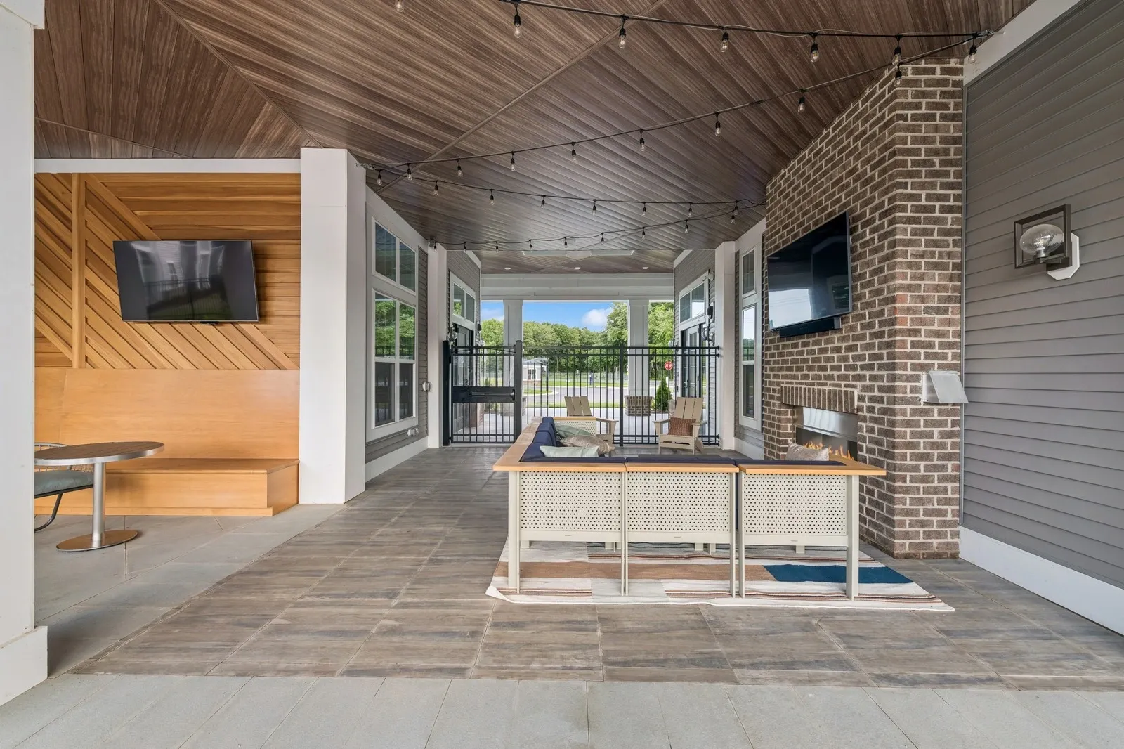 Covered outdoor lounge with brick fireplace, TVs, string lights, and seating.