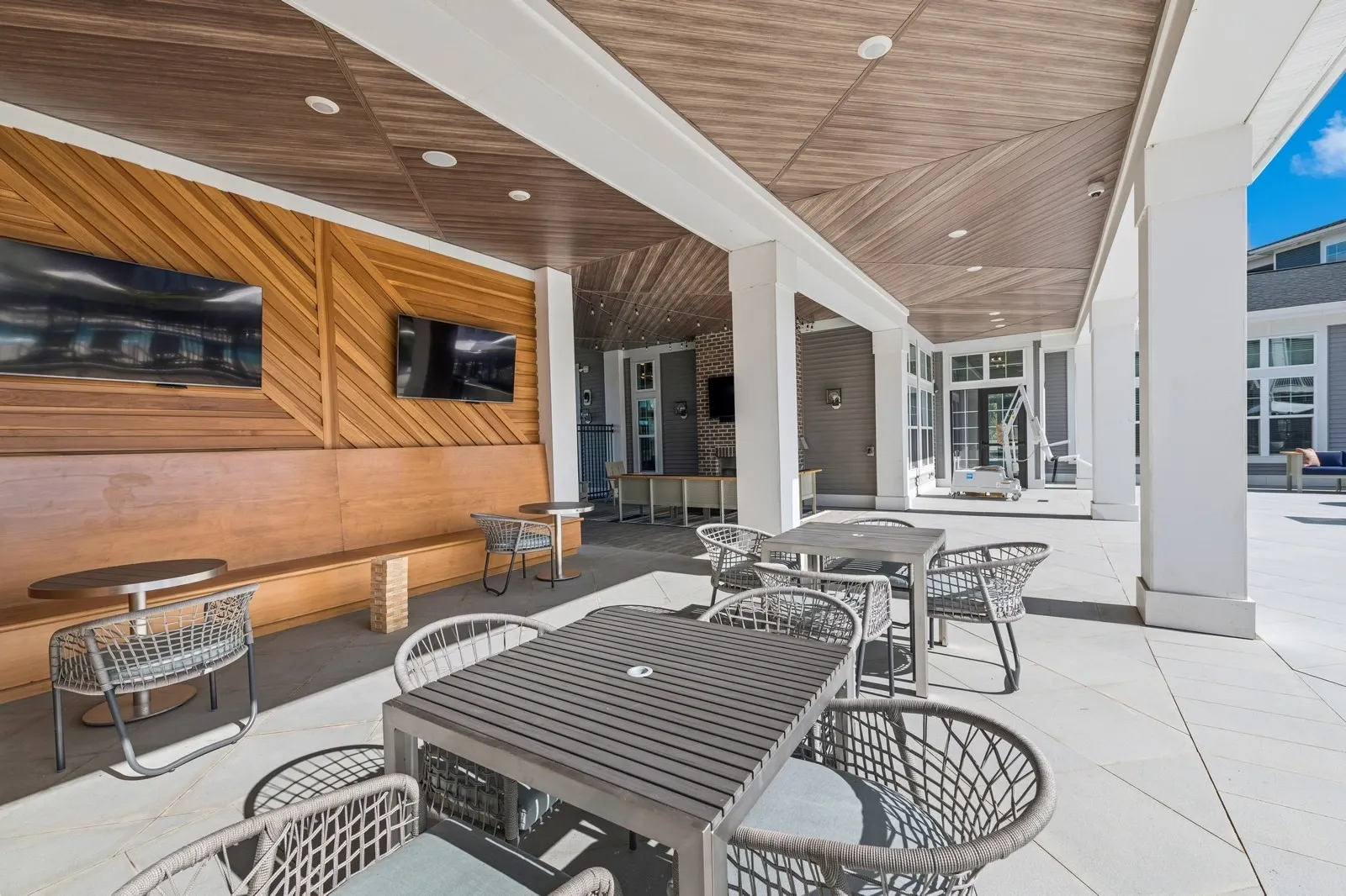 Outdoor community lounge with wooden wall, TVs, and metal tables under a covered patio.