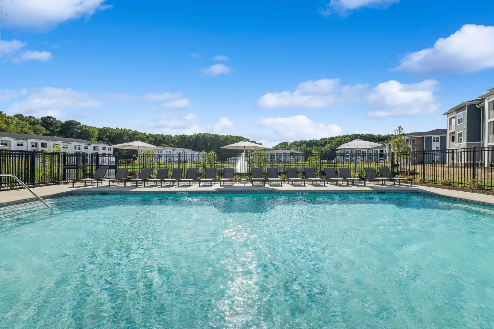 Outdoor community pool with a row of lounge chairs, umbrellas, and a black metal fence.
