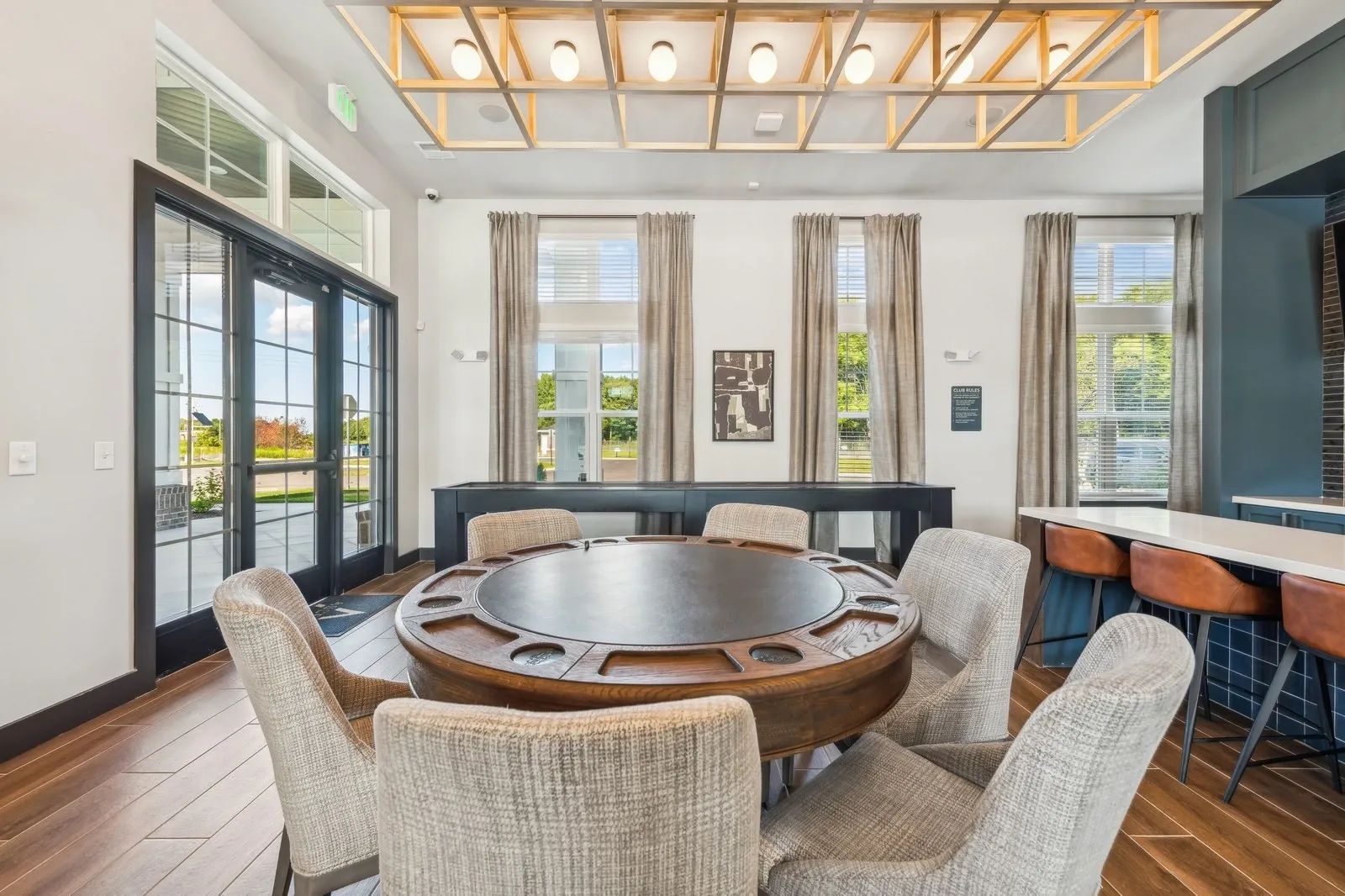 Communal lounge featuring a round wooden game table, beige upholstered chairs, and a bar-height coun…