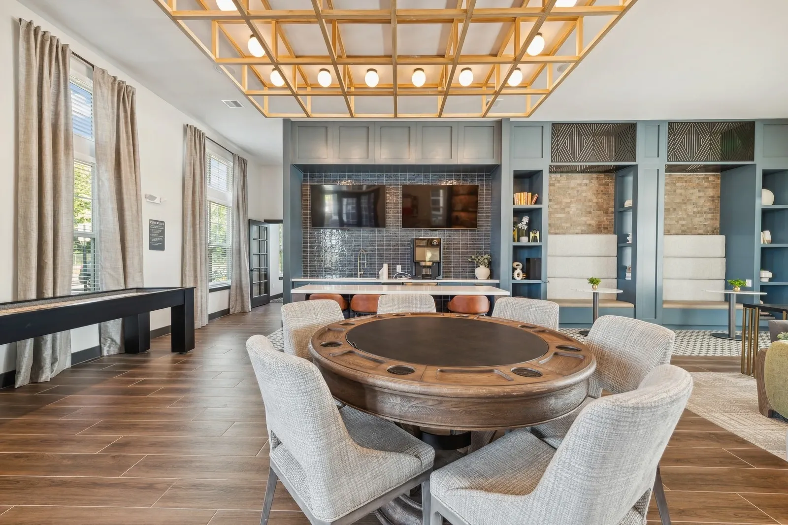 Community lounge with a round wooden table, beige upholstered chairs, and a kitchenette area.