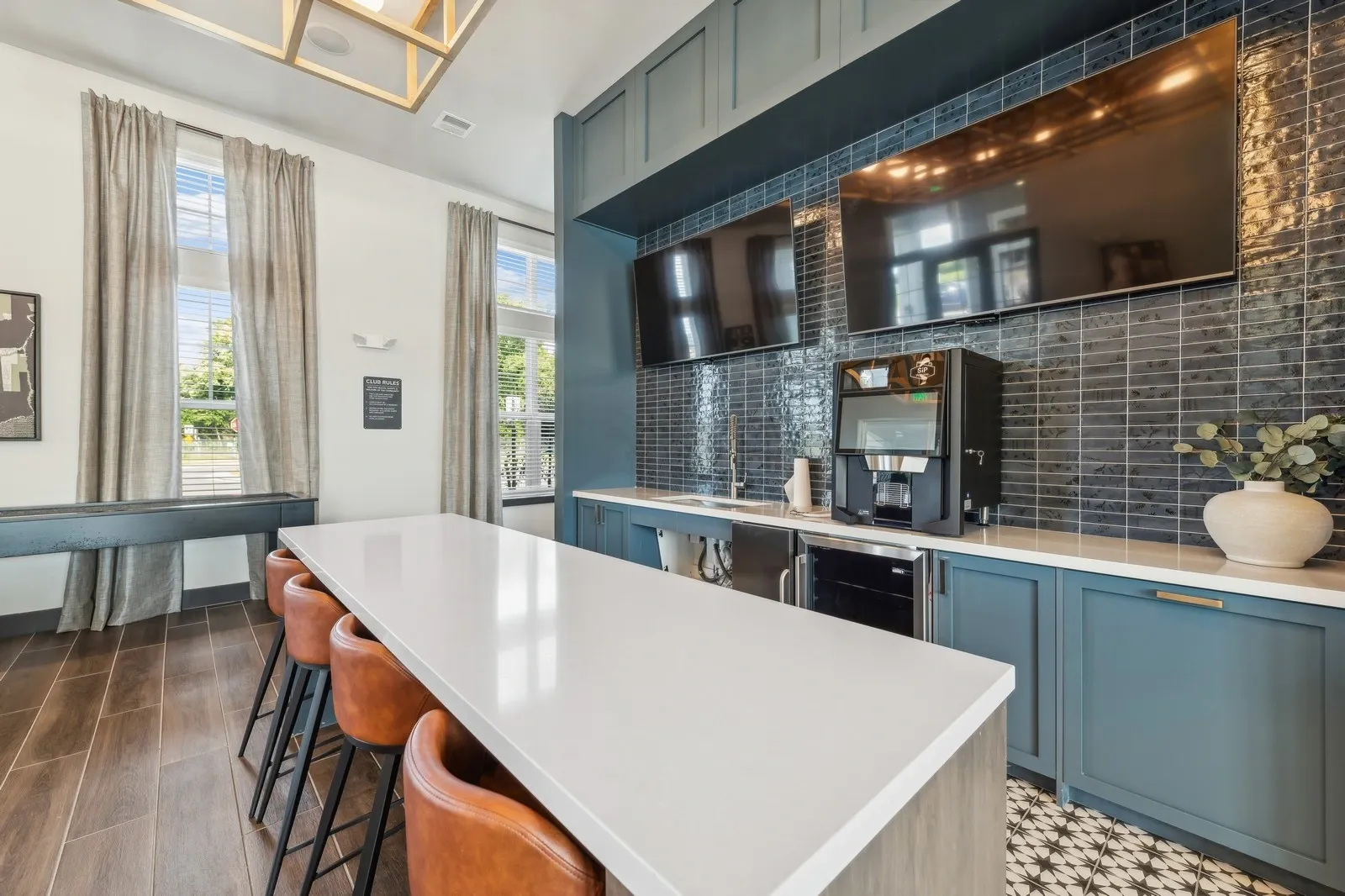 Community kitchen area with a white island, blue cabinets, tiled backsplash, and two wall-mounted TV…