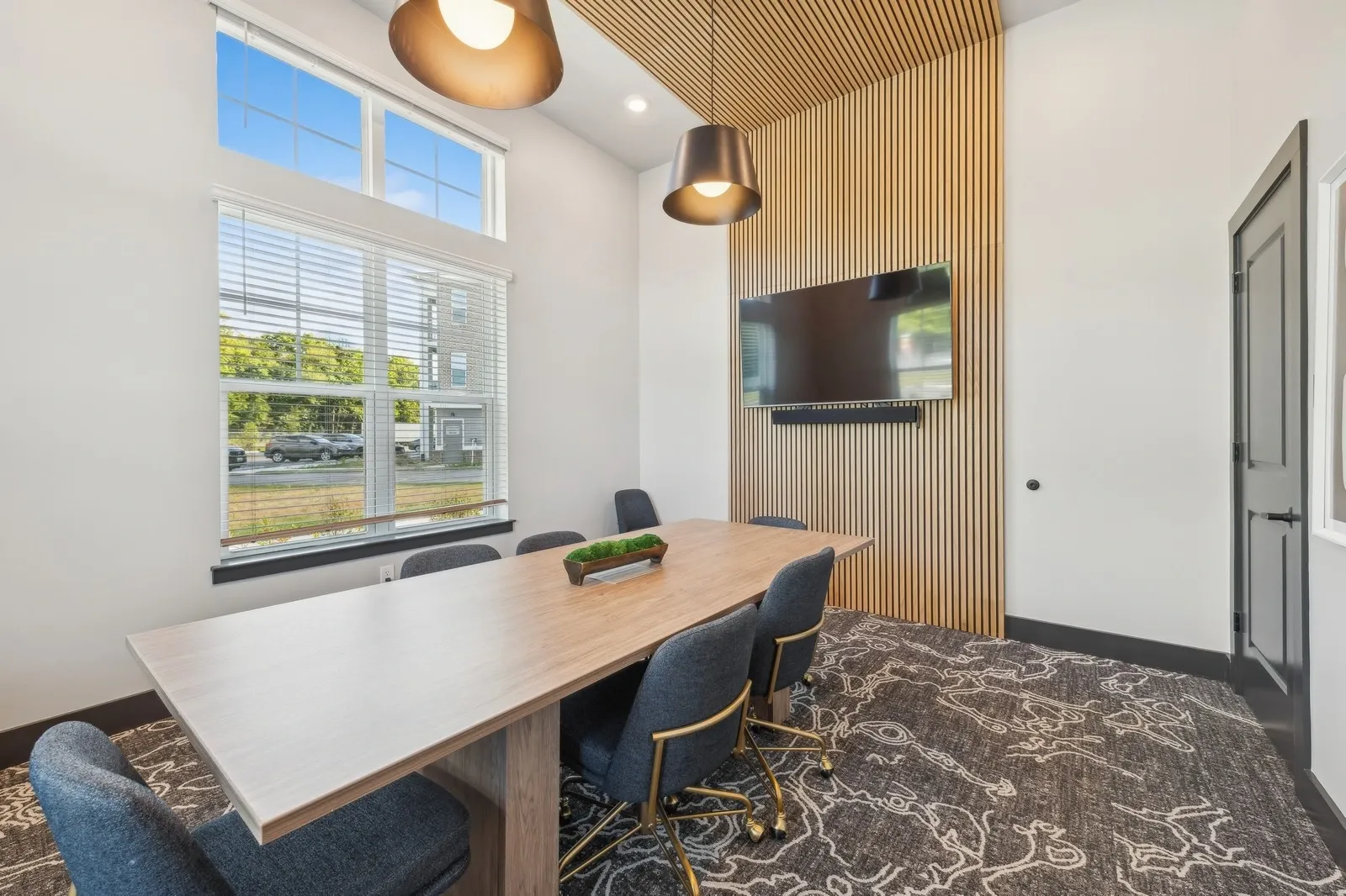 Modern conference room with wooden slat wall, mounted TV, long table, and blue chairs.