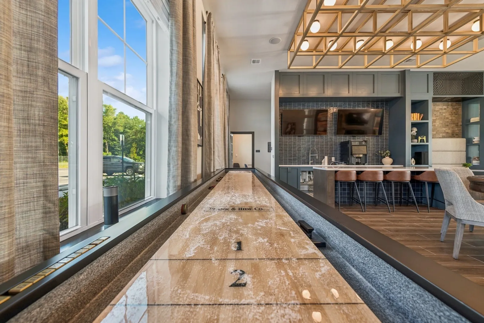 Indoor community lounge with a long shuffleboard table, windows, and a bar area.