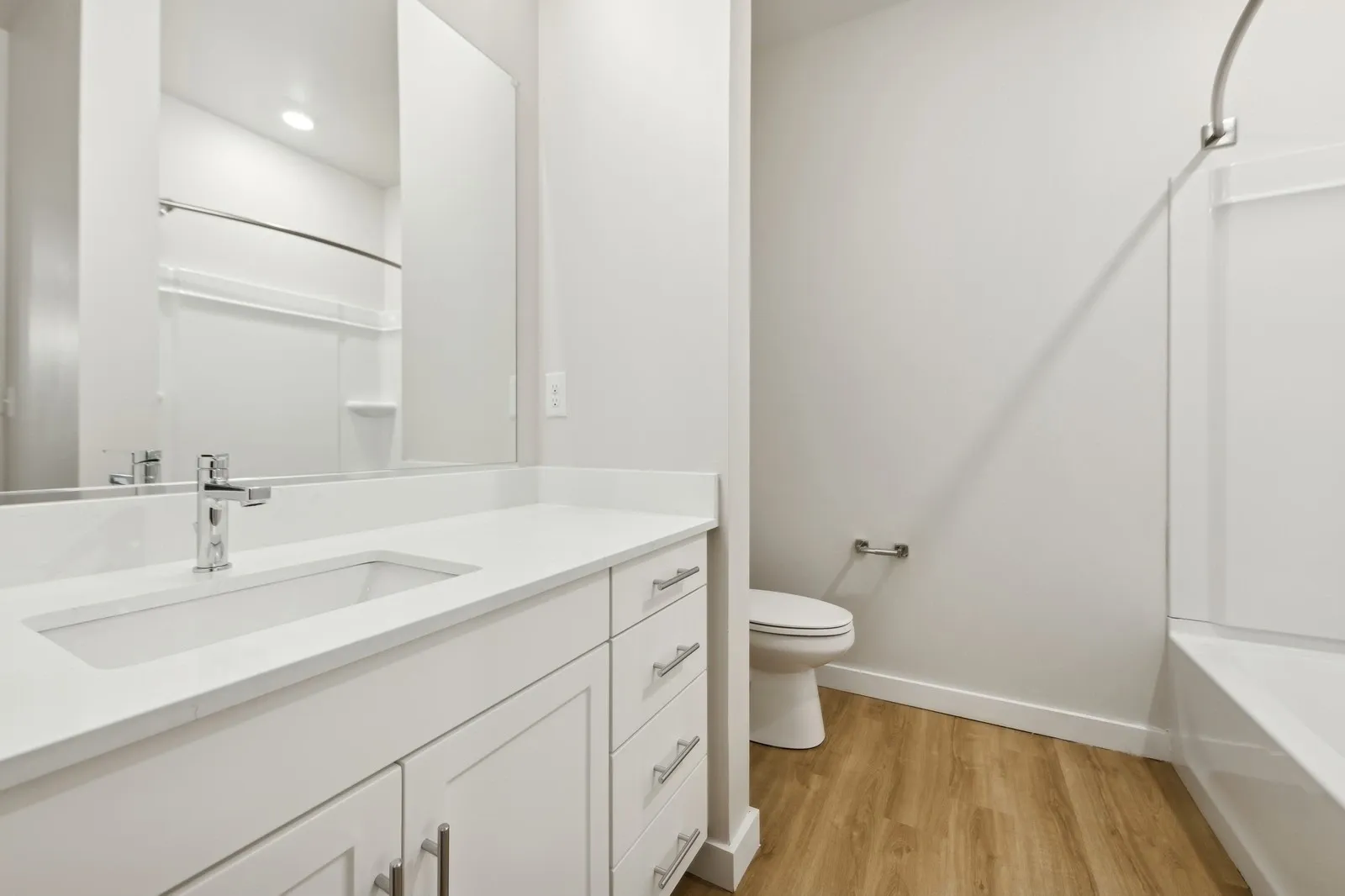 White bathroom with vanity, mirror, toilet, and tub/shower with wood-look flooring.