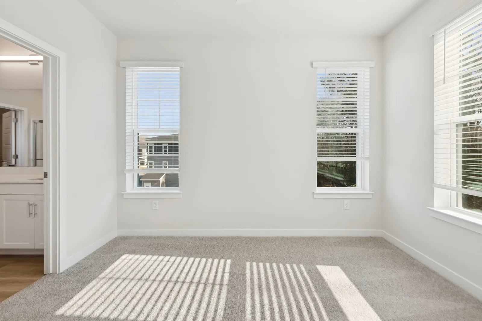 Bright empty bedroom with two windows, white walls, beige carpet, and an open doorway to a bathroom.