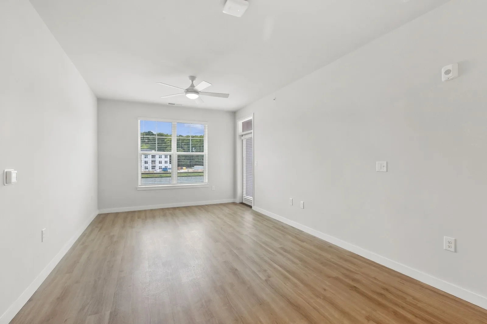 Empty living room with wood-like flooring, white walls, ceiling fan, and a large window