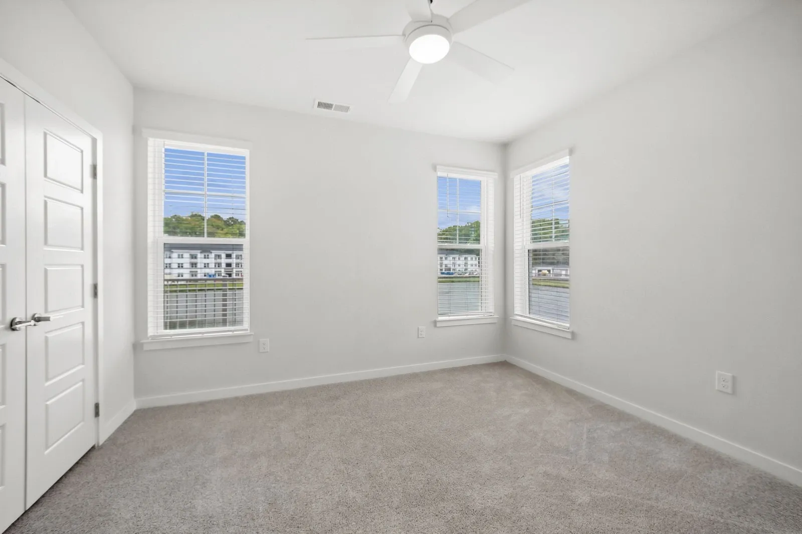 Bright, unfurnished bedroom with white walls, beige carpet, and a ceiling fan, featuring large windo…
