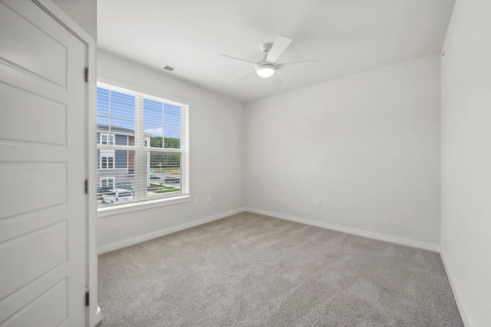 Bright empty bedroom with white walls, beige carpet, a large window with white blinds, and a ceiling…