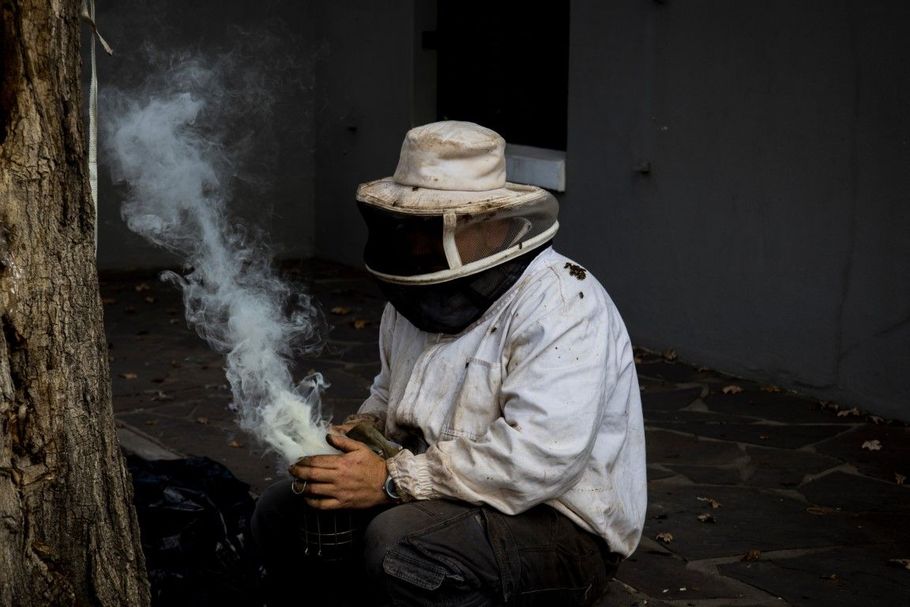 Person in a hat and face covering sitting and smoking beside a wall, with smoke rising in a dim room.