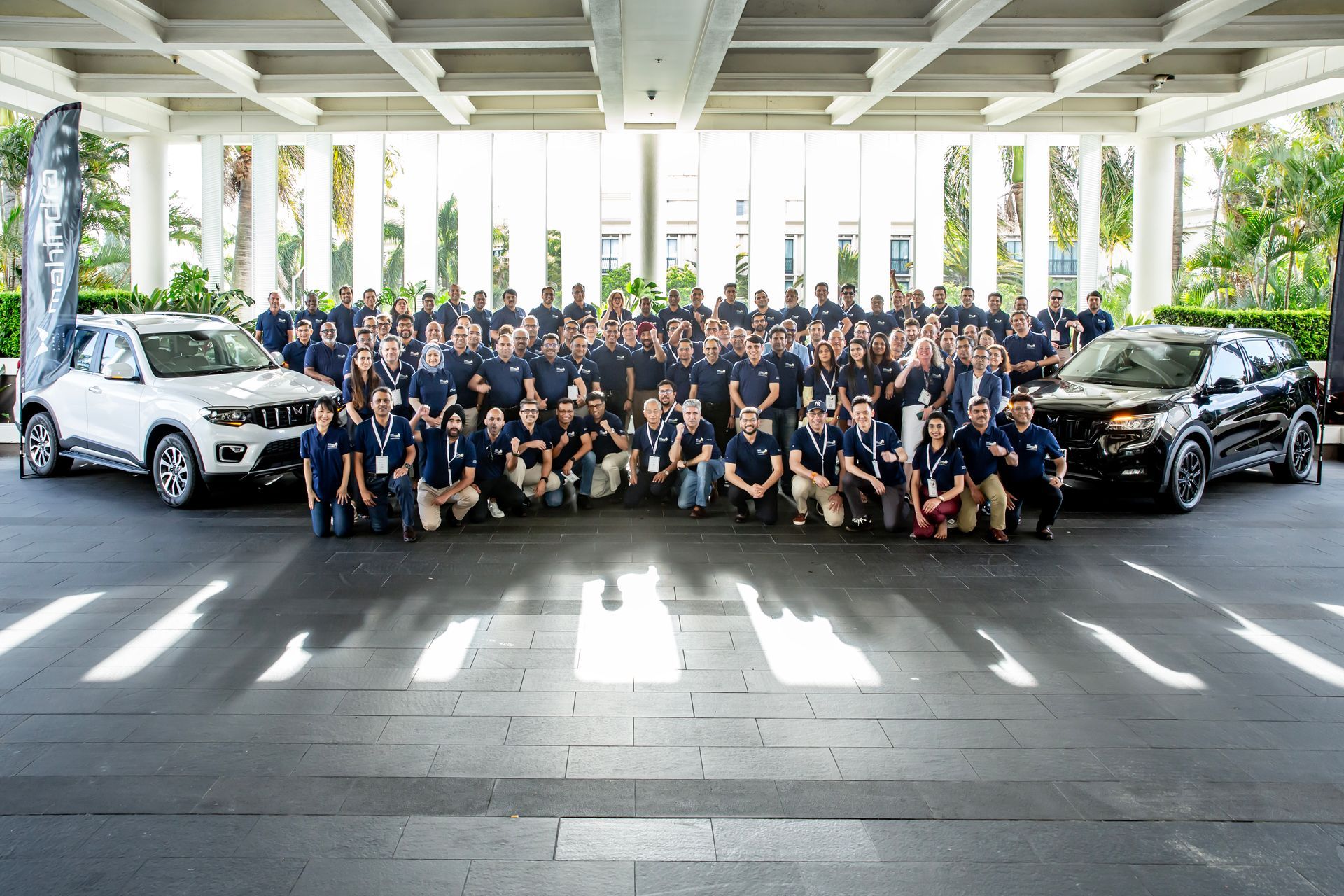 Large group of people posing with two SUVs under a building's portico. Most wear blue shirts.