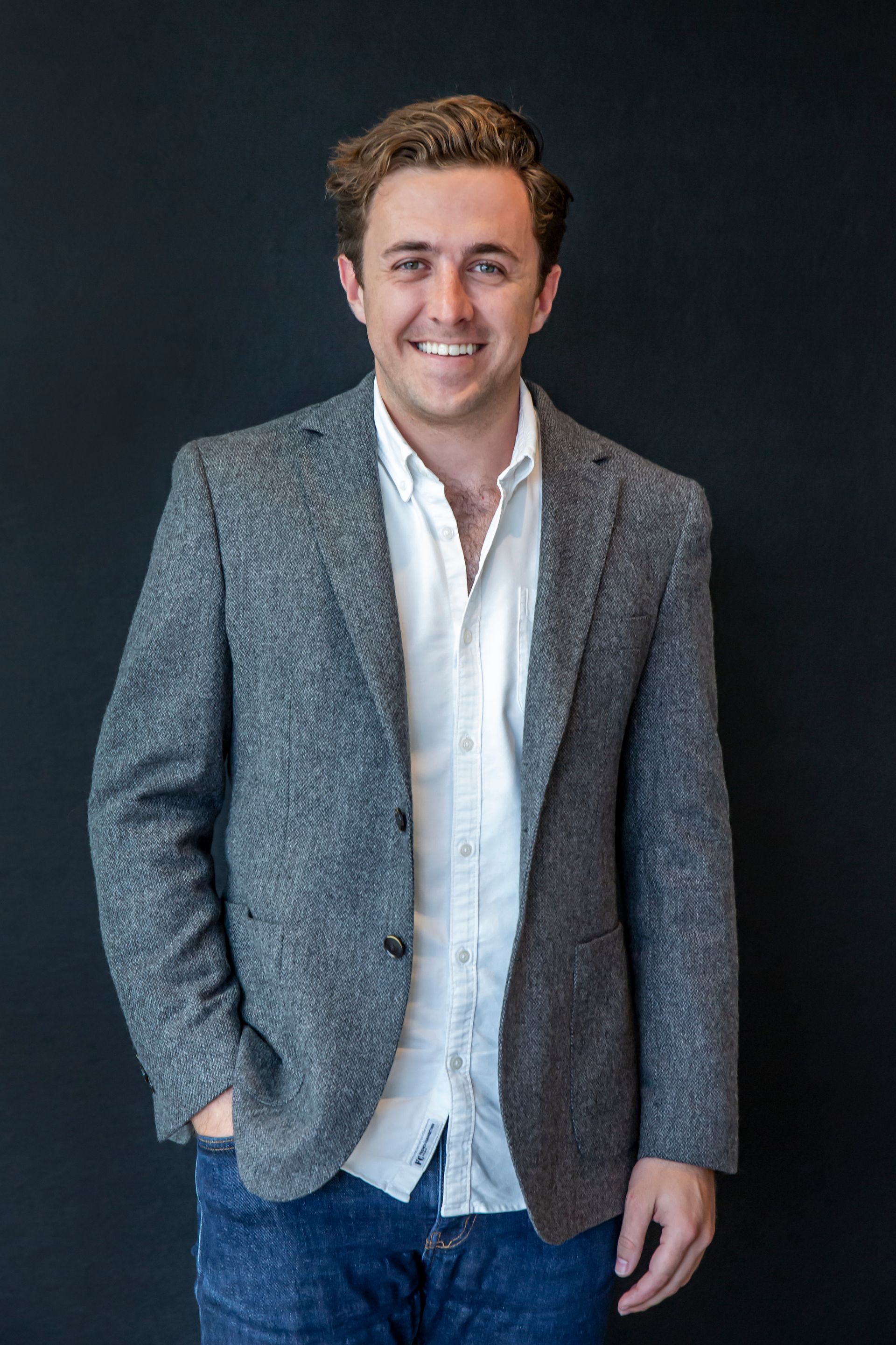 Young man in a gray blazer and white shirt smiles, one hand in pocket, against a black wall.