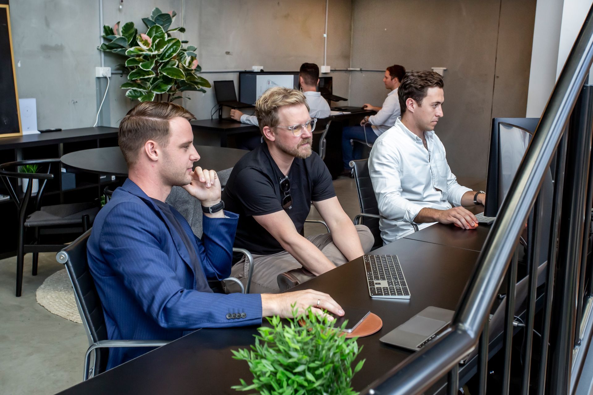 Three men in a modern office setting, working on computers; two looking at the screen intently.