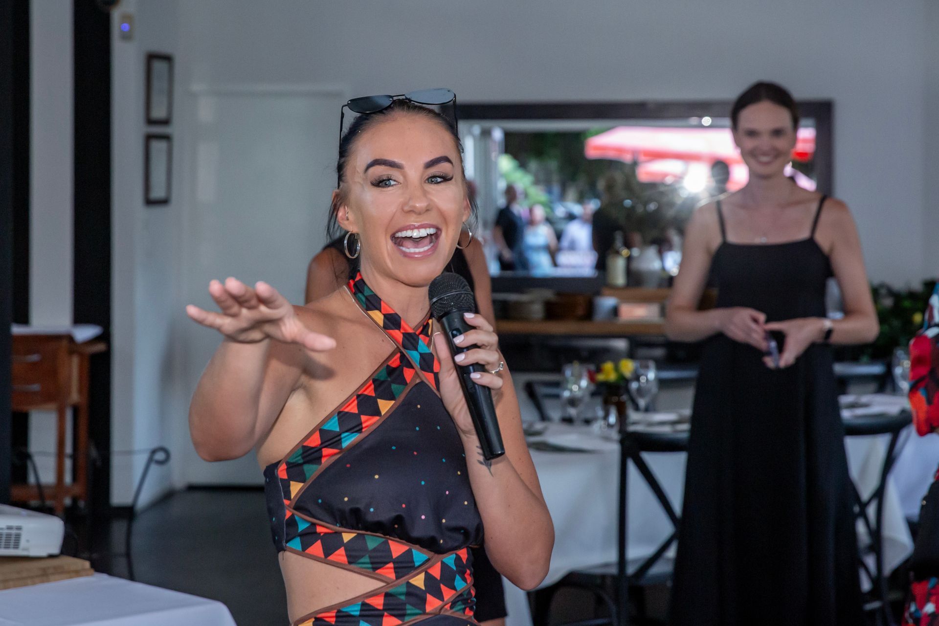 Woman with microphone gesturing enthusiastically at an event, another woman stands in the background.