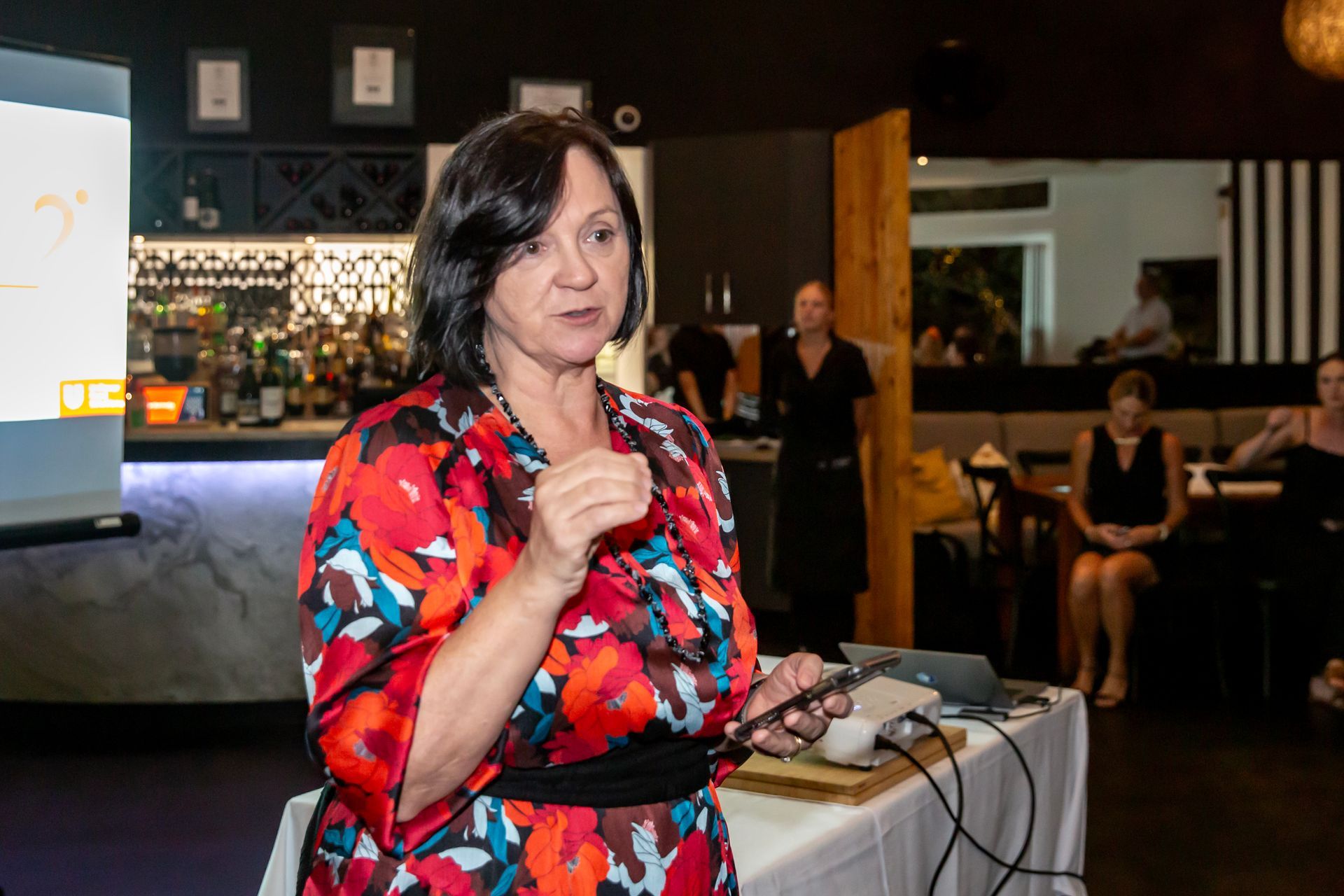 Woman giving a presentation in a restaurant, gesturing with a microphone; a screen and audience are visible.