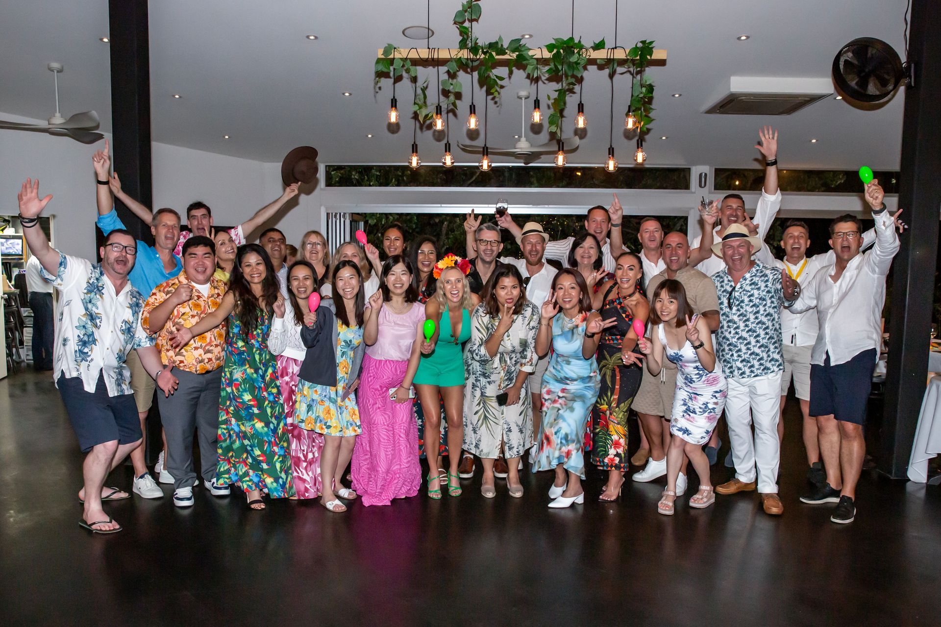 Group of people in tropical attire, smiling, raising hands at a party indoors with greenery. Photography by Andy Williams