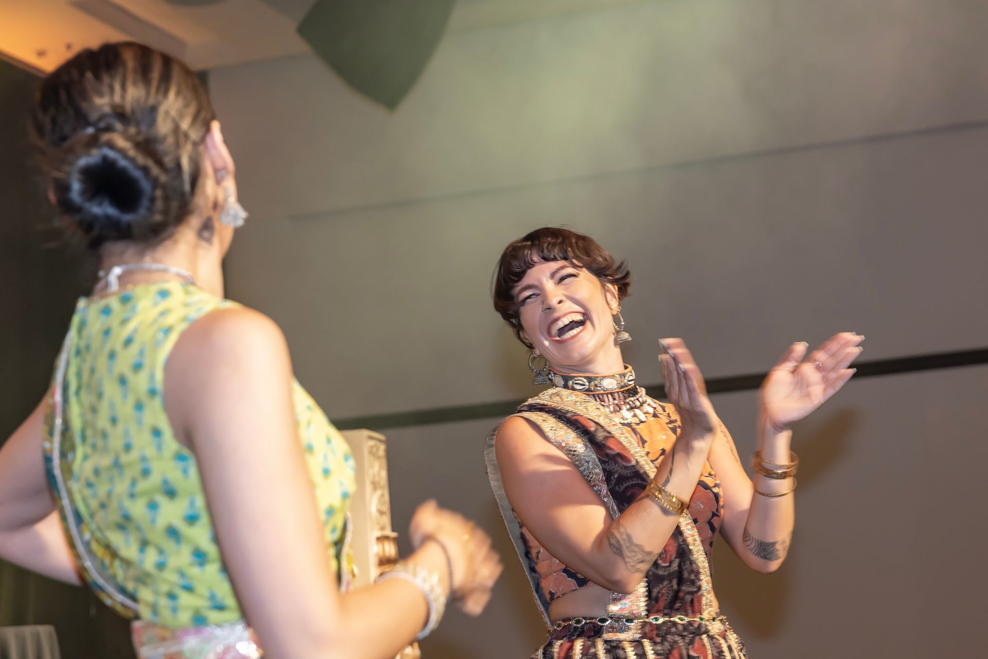 Two women in colorful outfits laugh and clap onstage, under bright lights.