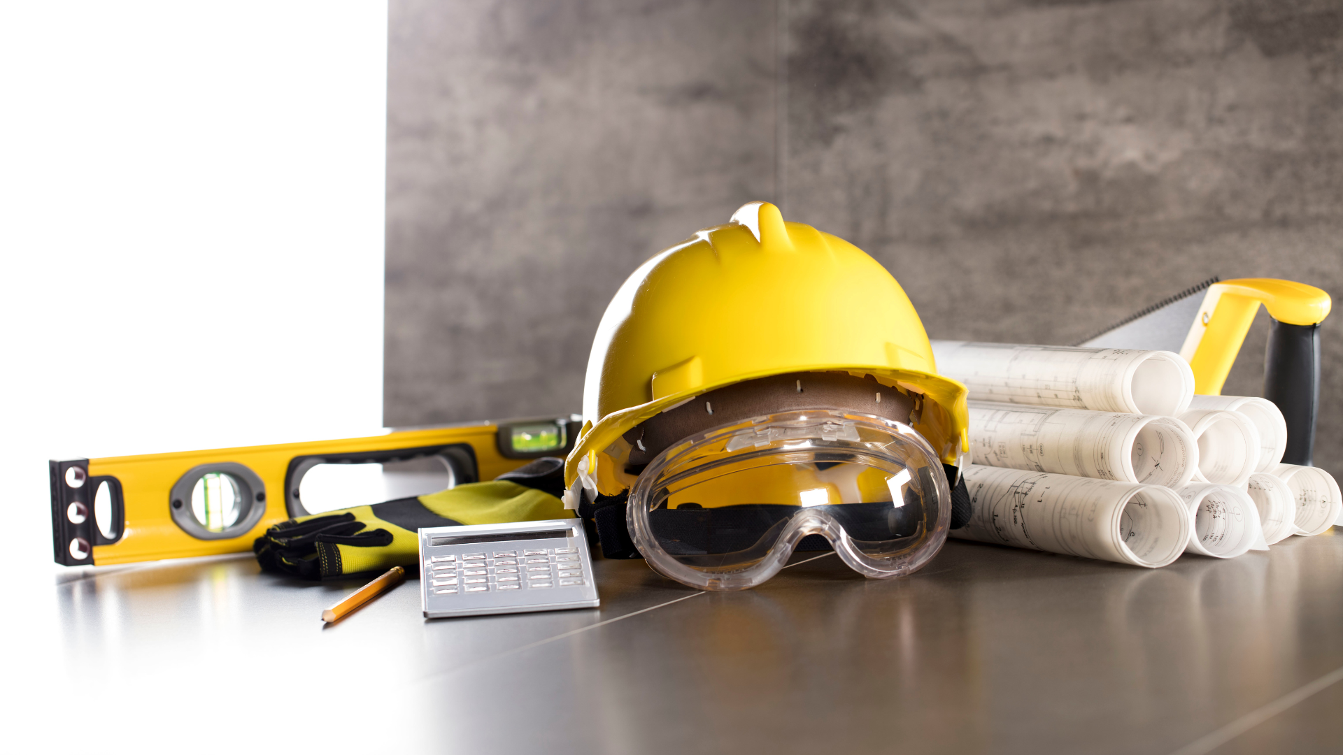 Construction tools: yellow hard hat, safety glasses, level, saw, blueprints on a metal surface.