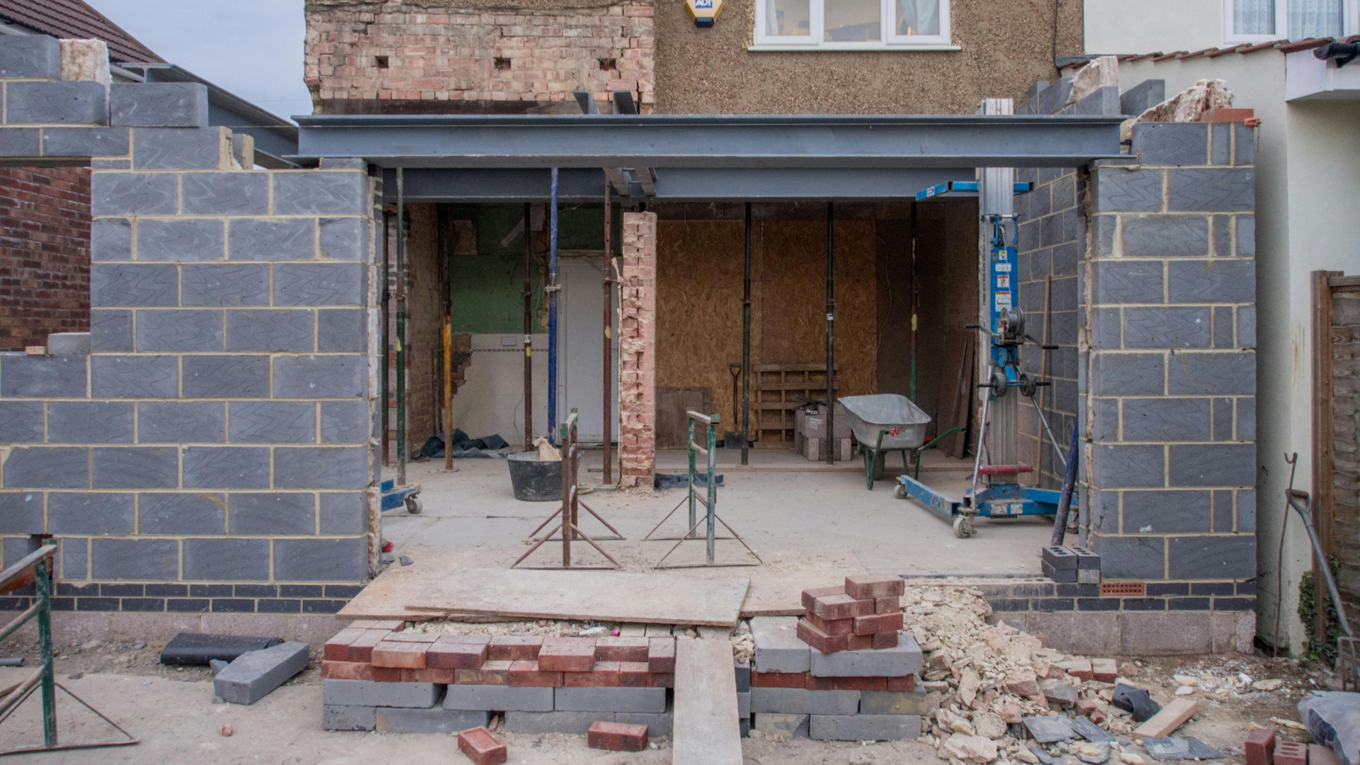 Construction site: bricklayers building a house extension, steel beam support, cinder block walls, brick debris.