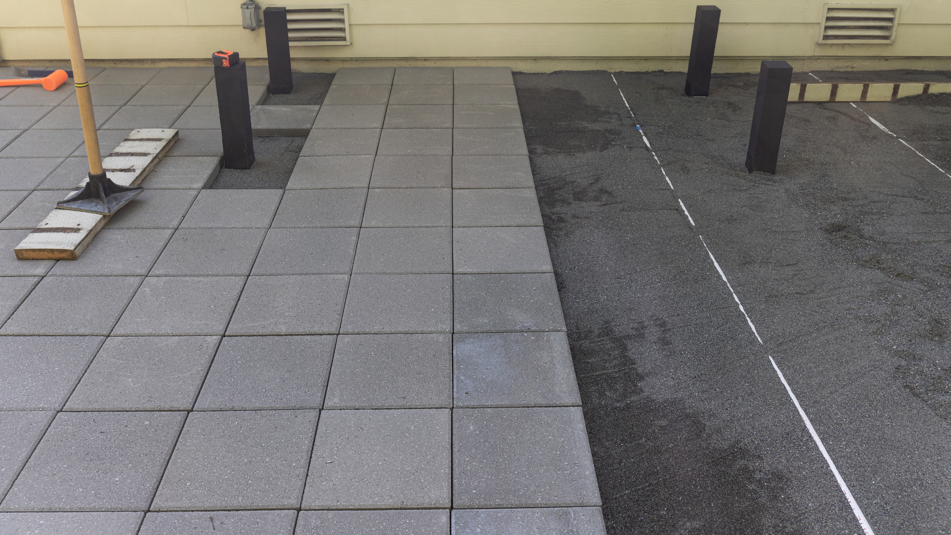 Patio construction: worker laying gray tiles on a gravel base, near black support posts.
