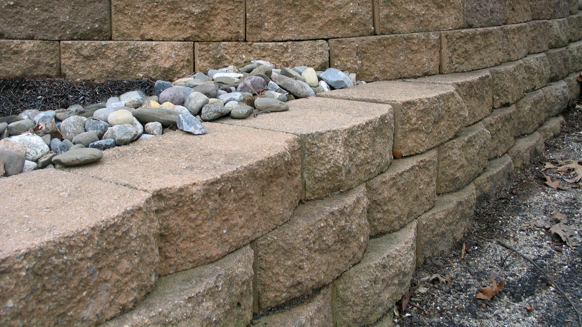 Tan retaining wall made of stone blocks with gravel and small rocks.