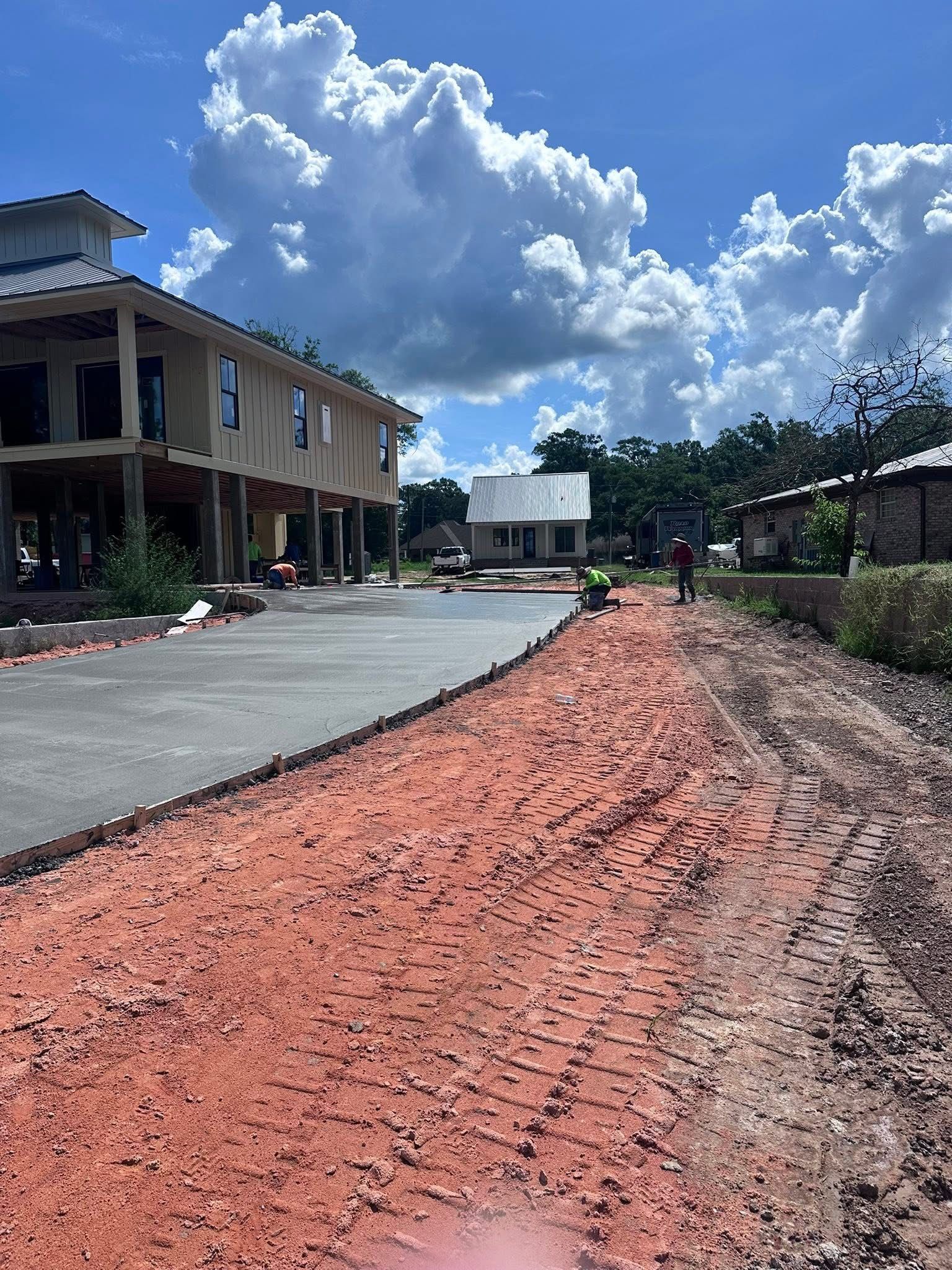 Red dirt roadside beside a paved drive, with a two-story building and bright cloudy sky.