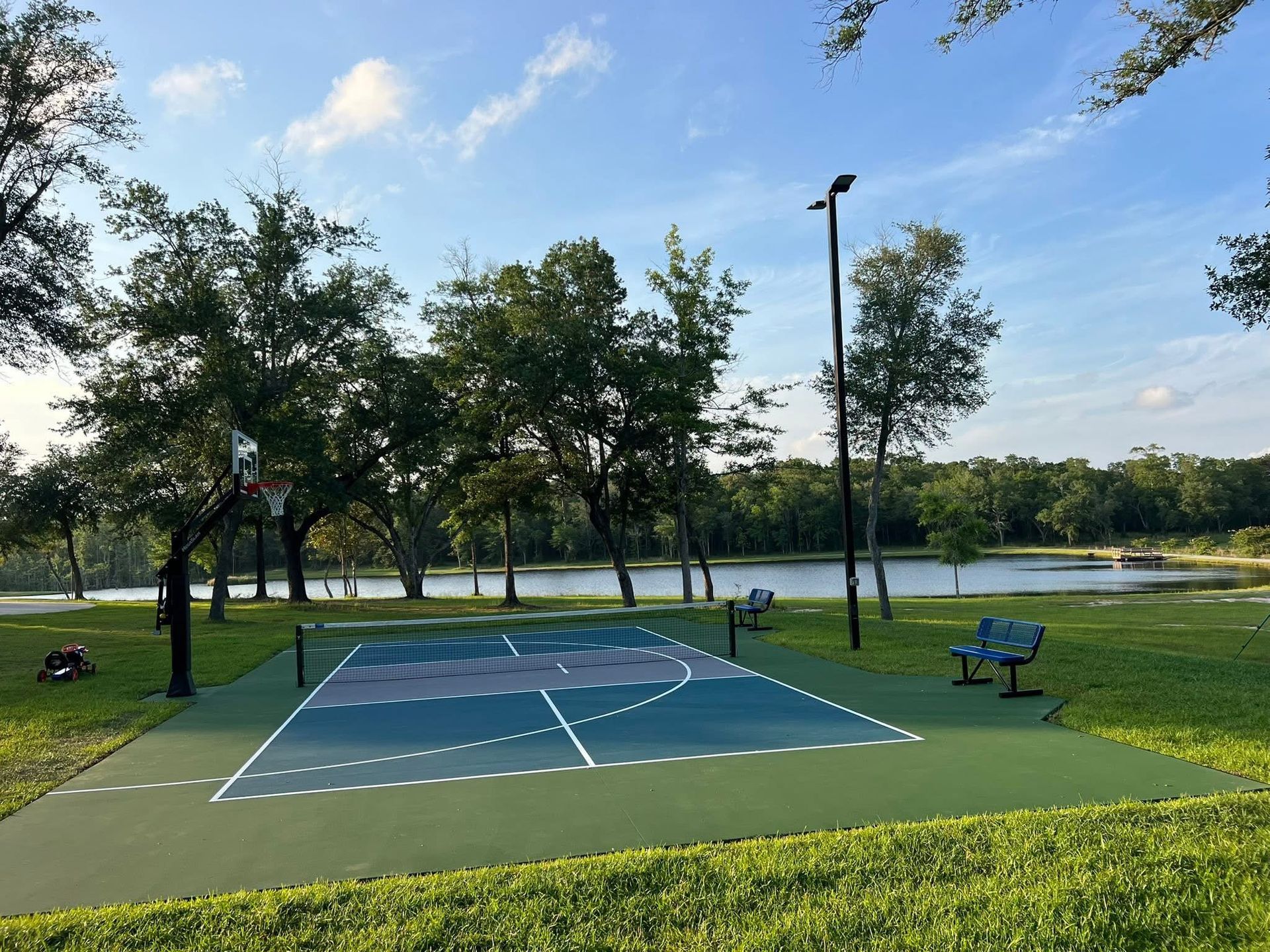 Outdoor pickleball court beside a lake, with trees, blue sky, and empty benches